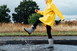© okrasiuk - Woman having fun on the street after the rain. Cropped woman wearing rain rubber boots and yellow raincoat walking into puddle with water splash and drops. Fall weather. Selective focus