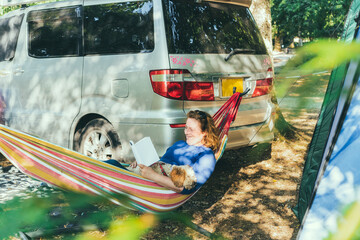 Adult woman relaxing and reading book while lying in hammock with cockapoo puppy near motorhome on camping trip. Female living on camper car with animals and travel the world. Caravan car Vacation