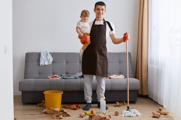 Wall Mural - Indoor shot of smiling satisfied father washing floor with mop at home and playing with his infant kid, doing domestic chores, cleaning apartment, looking at camera.