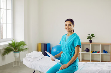 Wall Mural - Portrait of smiling female masseuse or doctor in uniform hold patient card pose in cabinet in hospital. Happy young woman physiotherapist or therapist in clinic. Healthcare and medicine concept.