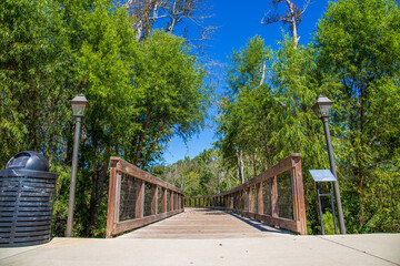 the entrance to a long winding brown wooden bridge with two black lamp posts surrounded by lush green trees and plants with blue sky at The Walk at Sandy Run in Warner Robins Georgia USA