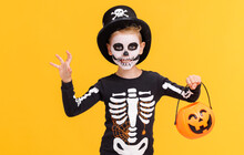 Happy Cheerful Boy In Skeleton Costume With  Pumpkin  Basket Celebrates Halloween And Scary Gesture  On Yellow Background