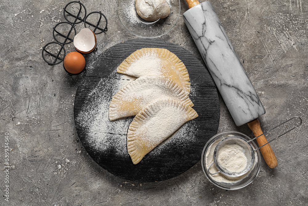 Board with raw chebureks, eggs, flour and rolling pin on grunge background