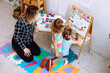 © Татьяна Волкова - Educator teach little children read and count while sitting on floor in playing room. Little girls arrange numbers and letters on magnetic board in kindergarten. Lesson, preschool, daycare