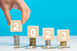© nazariykarkhut - Hand place wooden cube 2023 with stack of coins with blue background and copy space. Saving money and financial plan concept for investment in new year 2023.