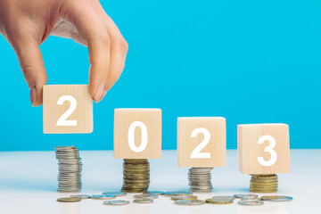 Hand place wooden cube 2023 with stack of coins with blue background and copy space. Saving money and financial plan concept for investment in new year 2023.