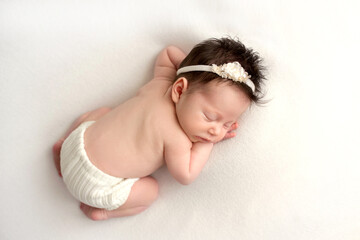 Top view of a newborn baby girl sleeping in white panties, with a white bandage her head on a white bed. Beautiful portrait of a little girl 7 days, one week. Professional photography in macro studio.