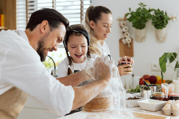 Close-up shot happy father sieving flour from bag on wooden surface curious daughter helping watching process. In background mother with ponytail opening sauce for homemade pizza.