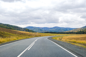 Mountain roadway. Road in autumn forest in Siberia. Trees with orange foliage. Landscape with empty asphalt road through woodland, blue sky, high rocks in fall. Travel concept. 