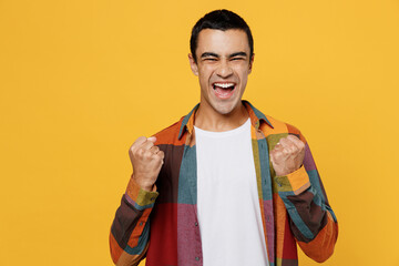 Young fun middle eastern man 20s he wear casual shirt white t-shirt doing winner gesture celebrate clenching fists say yes isolated on plain yellow background studio portrait People lifestyle concept.