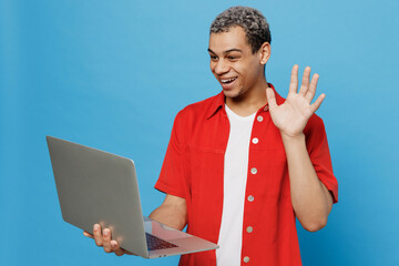 Wall Mural - Young smiling happy fun IT man of African American ethnicity 20s he wear red shirt hold use work on laptop pc computer waving hand get video call isolated on plain pastel light blue cyan background.