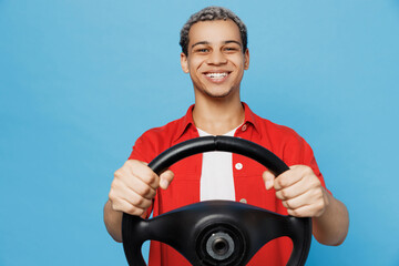 Young cheerful happy fun man of African American ethnicity 20s he wearing red shirt hold steering wheel pretend driving isolated on plain pastel light blue cyan background. People lifestyle concept.