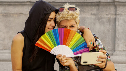 Wall Mural - a non-binary persons with a rainbow hand fan looking at mobile. Gay Pride's day