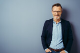 Fototapeta Na drzwi - Confident middle-aged man in blue blazer and blue shirt, wearing glasses, standing against plain blue background with copy space and looking at camera