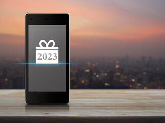 Gift box happy new year 2023 flat icon on modern smart mobile phone screen on wooden table over blur of cityscape on warm light sundown, Business happy new year 2023 shop online concept