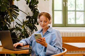 Wall Mural - Young white businesswoman using headphones and cellphone in cafe