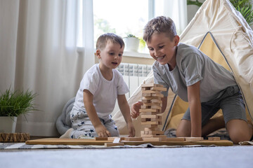 Two male children playing Jenga hardwood bricks tower construction childish room with linen wigwam