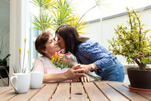 Ethnic Woman Kissing Elderly Mother And Presenting Flowers On Terrace