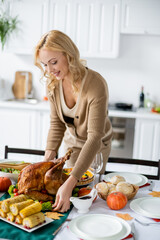 Wall Mural - happy blonde woman serving table with roasted turkey near grilled corn and buns for festive thanksgiving dinner