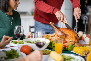 Wall Mural - Cropped view of senior man cutting delicious thanksgiving turkey near women and wine at home