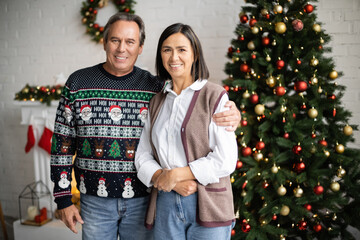 Wall Mural - smiling man in sweater with festive pattern hugging multiracial wife near blurred christmas tree