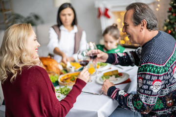 Wall Mural - cheerful relatives clinking glasses of red wine near delicious christmas dinner and blurred family