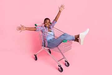 Wall Mural - Full size portrait of overjoyed crazy lady sit shopping trolley raise hands isolated on pink color background