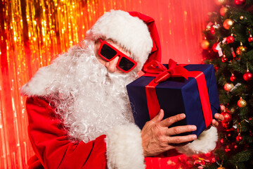 Canvas Print - Father christmas in sunglasses and costume holding gift box during party near pine tree.