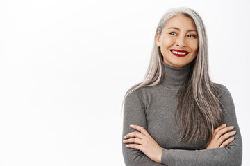 Portrait of beautiful, healthy smiling middle aged asian woman, cross arms on chest, looking confident and happy, standing over white background