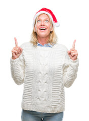 Poster - Middle age blonde woman wearing christmas hat over isolated background amazed and surprised looking up and pointing with fingers and raised arms.