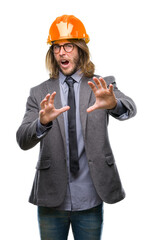 Poster - Young handsome architec man with long hair wearing safety helmet over isolated background afraid and terrified with fear expression stop gesture with hands, shouting in shock. Panic concept.