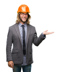 Sticker - Young handsome architec man with long hair wearing safety helmet over isolated background smiling cheerful presenting and pointing with palm of hand looking at the camera.