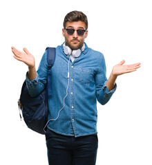 Young handsome tourist man wearing headphones and backpack over isolated background clueless and confused expression with arms and hands raised. Doubt concept.