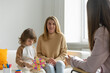 Young cheerful mother at a reception at a child family psychologist with her little son. Little boy with long haircut plays educational toys. Blurred foreground.