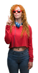 Sticker - Young redhead woman wearing headphones surprised with an idea or question pointing finger with happy face, number one