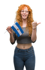 Sticker - Young redhead woman holding flag of Argentina pointing and showing with thumb up to the side with happy face smiling