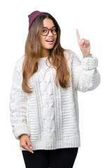 Canvas Print - Young beautiful brunette hipster woman wearing glasses and winter hat over isolated background pointing finger up with successful idea. Exited and happy. Number one.