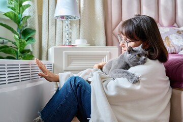 Wall Mural - Woman sitting with cat under blanket warming near an electric heater