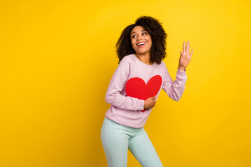 Wall Mural - Photo of young excited woman waving arm hello look empty space hold heart postcard isolated over yellow color background