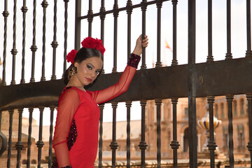 Wall Mural - Portrait of young teenage woman in red dance suit with red carnations in her hair doing flamenco poses holding on to a fence. Flamenco concept, dance, art, typical Spanish dance.