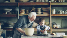Curious Little Boy Is Learning Pottery From His Experienced Grandfather In Small Home Studio. Child Is Forming Clay To Make Pot On Potter's Wheel, His Granddad Is Helping Him.