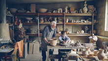 Experienced Potter Loving Prandparent Is Teaching His Young Grandchild Pottery In Small Home Workshop. Boy Is Working With Throwing Wheel, Grandpa Is Talking To Him.