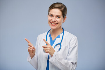 Wall Mural - Smiling doctor woman in white lab coat pointing with two fingers at left side, isolated portrait.