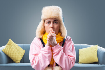 Portrait of young woman in a hat and yellow scarf is breathing, trying to keep warm. Gray background with sofa. The concept of energy crisis and winter season