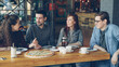 © silverkblack - Cheerful young friends are talking and gesturing sharing news while sitting at table in modern cafe together. Big pizza, cups and plates, tables and chairs are visible.