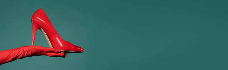 cropped view of woman in red glove holding high heeled shoe on green background, banner.