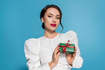 Wall Mural - brunette young woman in white blouse holding wrapped present and pooing at camera isolated on blue.