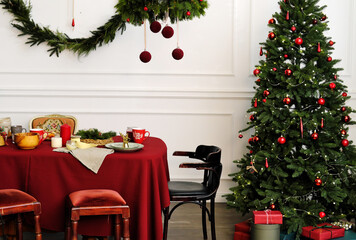 Canvas Print - Christmas table setting. On the table covered with a red tablecloth are plates, cups and festive decor. Christmas tree stands next to the table