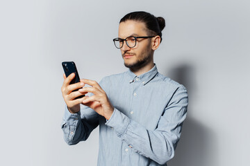 Wall Mural - Studio portrait of young man using smartphone on white background, wearing blue shirt and eyeglasses.