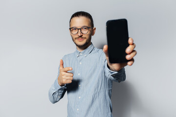 Wall Mural - Studio portrait of young man showing smartphone, pointing index finger on gadget, on white background. Wearing blue shirt and eyeglasses.